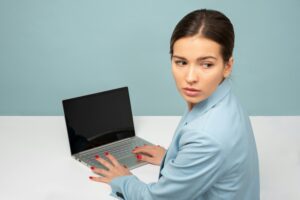 A woman in a blue blazer sits at a desk with a laptop, looking over her shoulder, vigilant against any signs of bullying or harassment in the office environment.