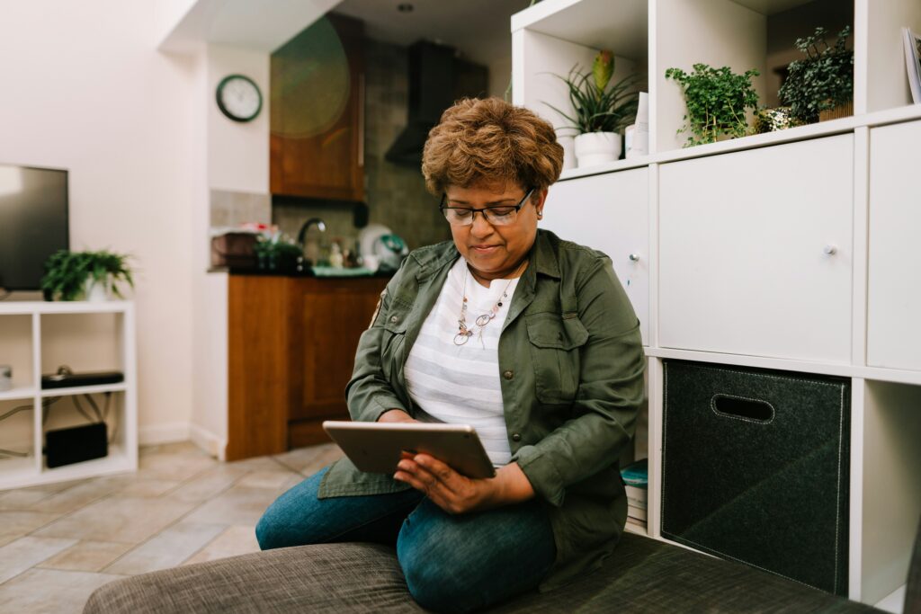 A person with bad posture is sitting on a couch in a living room, using a tablet. The room features white storage shelves and plants.