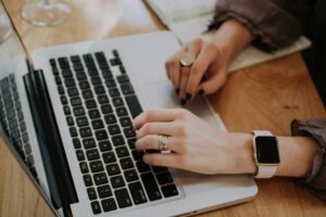 A person, deep in her-alysis, with rings and a smartwatch, types on a laptop, while a notebook lies nearby on the wooden table.