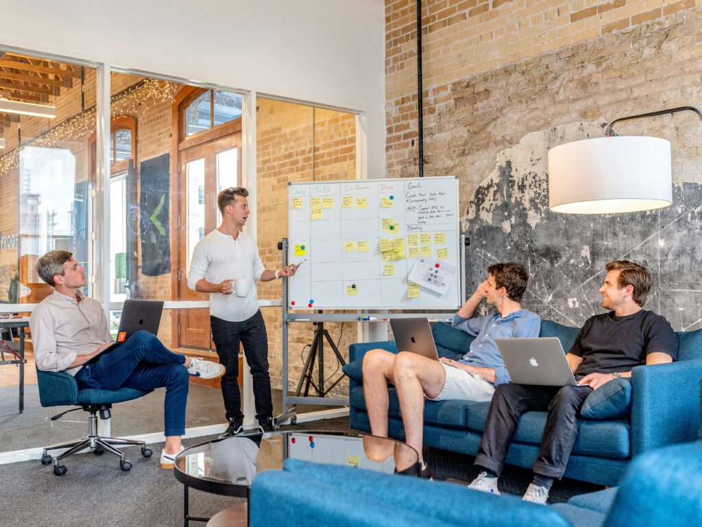 Four people in a casual meeting room discuss strategies for National Stress Awareness Week, pointing at a whiteboard filled with notes and using laptops.