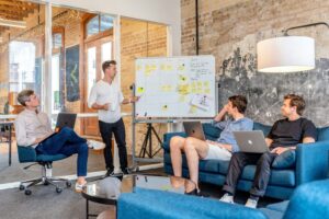 Four people in a casual meeting room discuss strategies for National Stress Awareness Week, pointing at a whiteboard filled with notes and using laptops.