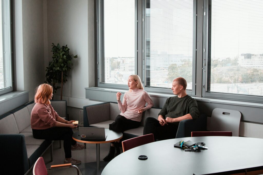 Three people sit in an office lounge, discussing the benefits of natural light streaming through large windows. A laptop rests on the table, and a lush plant adds a touch of green in the corner.