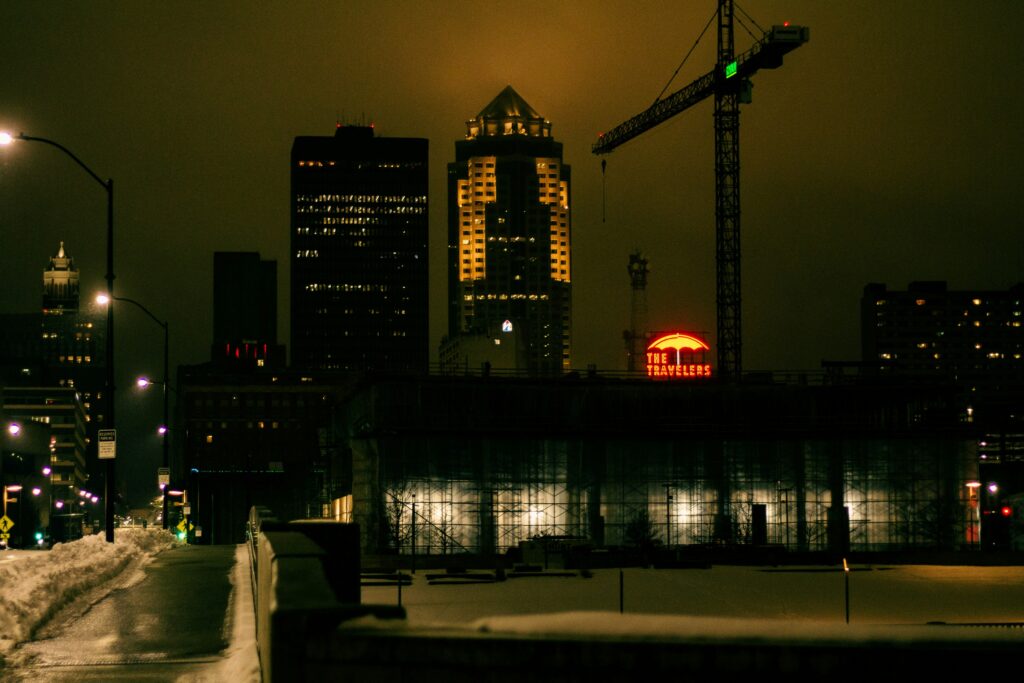 On a snowy night cityscape, illuminated buildings stand tall alongside a lone crane. A "The Travelers" sign flickers against the dark sky, while streetlights cast a melancholic glow on the path below, echoing whispers of solitude and depression in the frosty air.