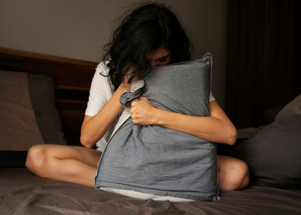 A person sits on a bed, battling insomnia, leaning into a gray pillow with their head partially obscured.