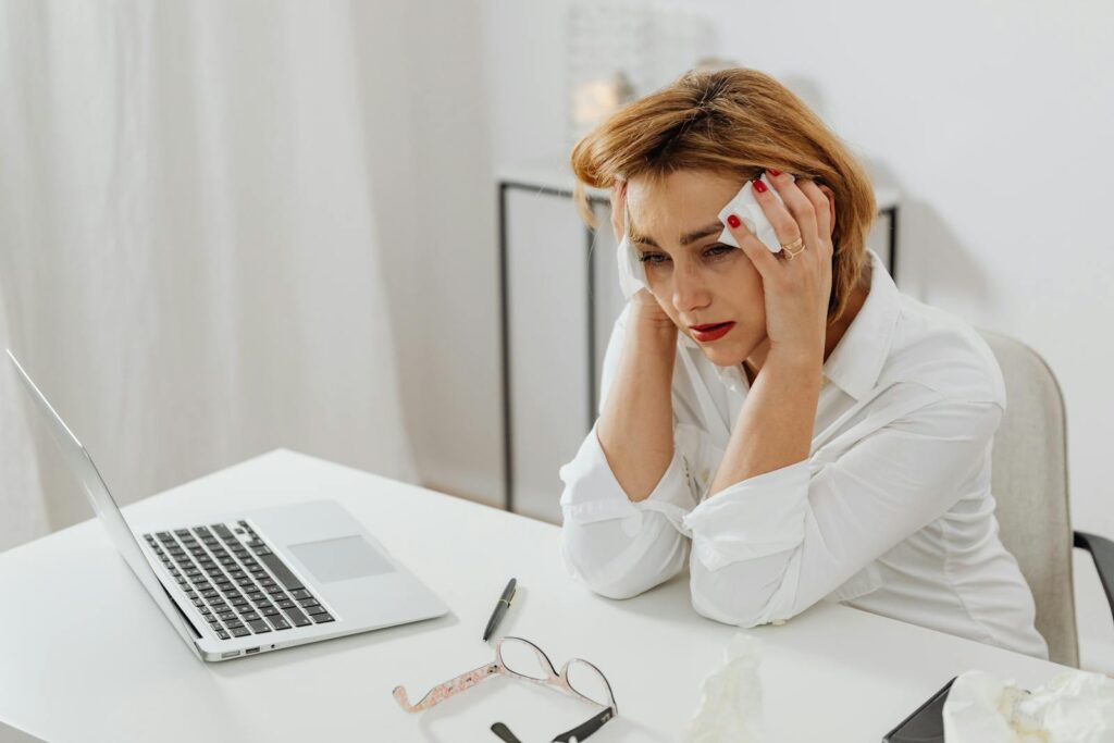 A woman in distress at her office desk, surrounded by tissues and a laptop.