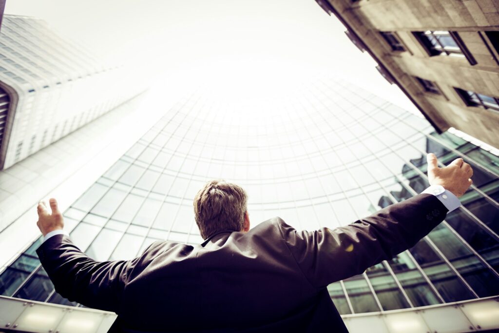 A man in a suit stands in front of tall office buildings with his hands outstreched.