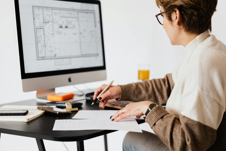 Woman designing architectural blueprints using desktop computer in a modern office setting.