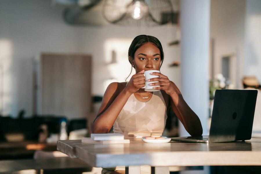 A stylish woman drinking coffee and working on a laptop indoors, creating a relaxed atmosphere.