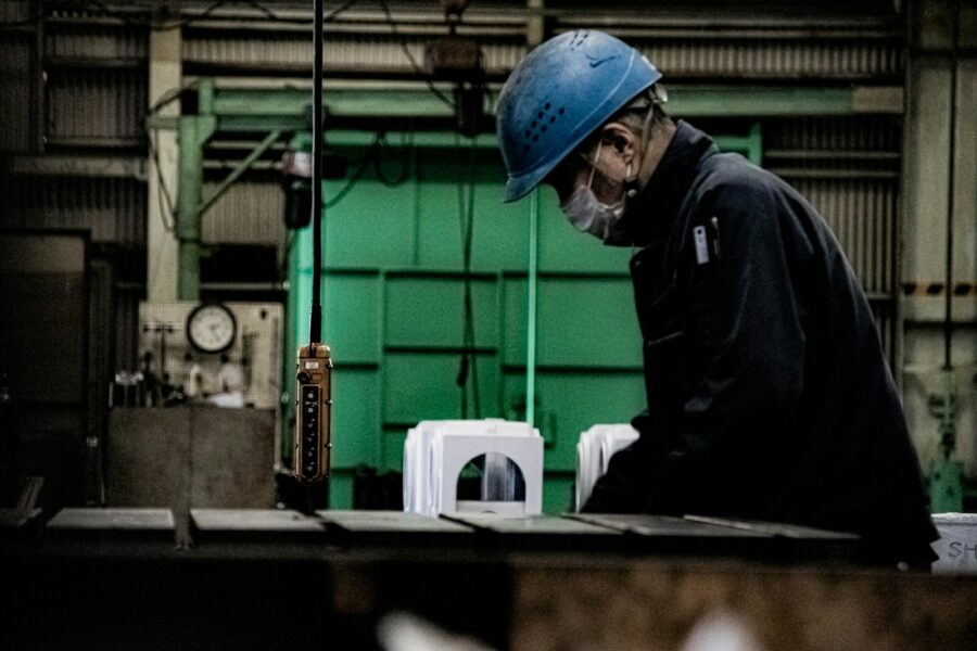 A man in blue jacket and blue cap standing in front of brown wooden table in a factory.