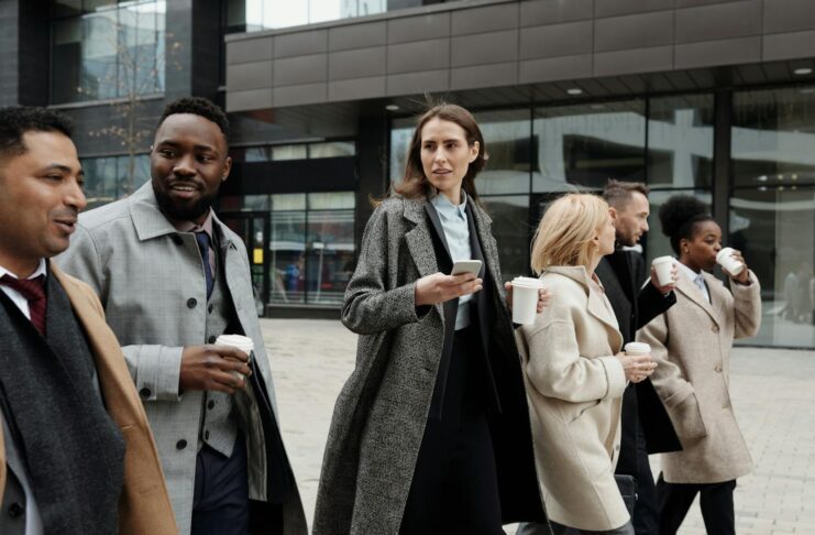 A diverse group of business professionals walking outside a modern building, enjoying coffee and conversation.
