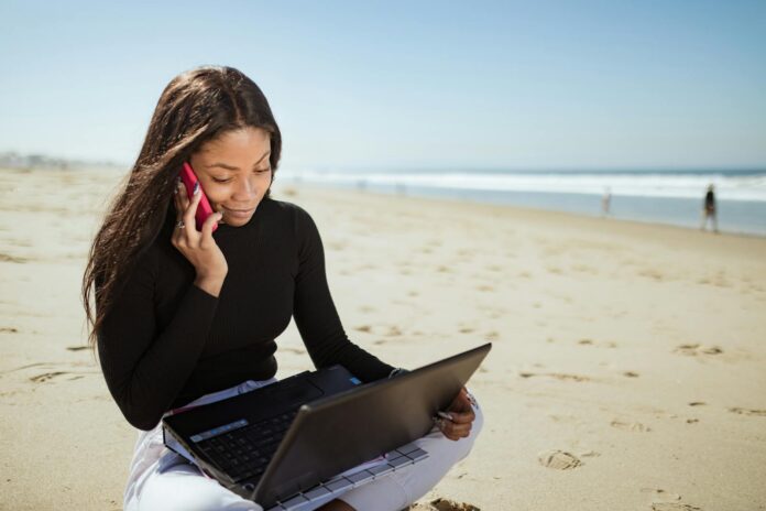 Woman multitasks on a beach, talking on phone while using laptop. Ideal for remote work imagery.