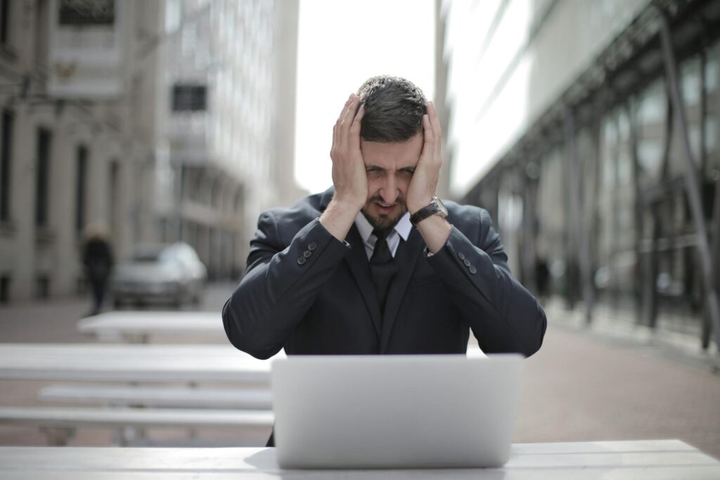 A businessman in a suit looks stressed, sitting outdoors with a laptop in a city environment.