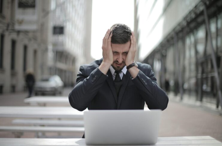 A businessman in a suit looks stressed, sitting outdoors with a laptop in a city environment.