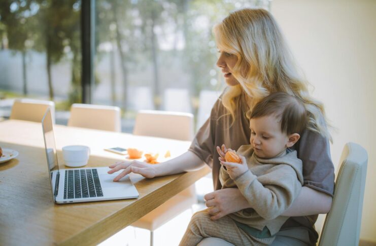 A mother works on a laptop at home while holding her baby, showcasing remote working and parenting.