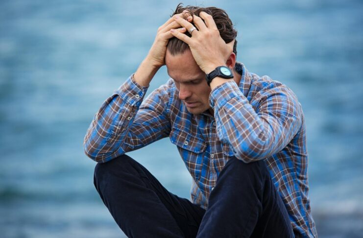 A man in a plaid shirt sits by the water looking distressed, symbolizing stress.