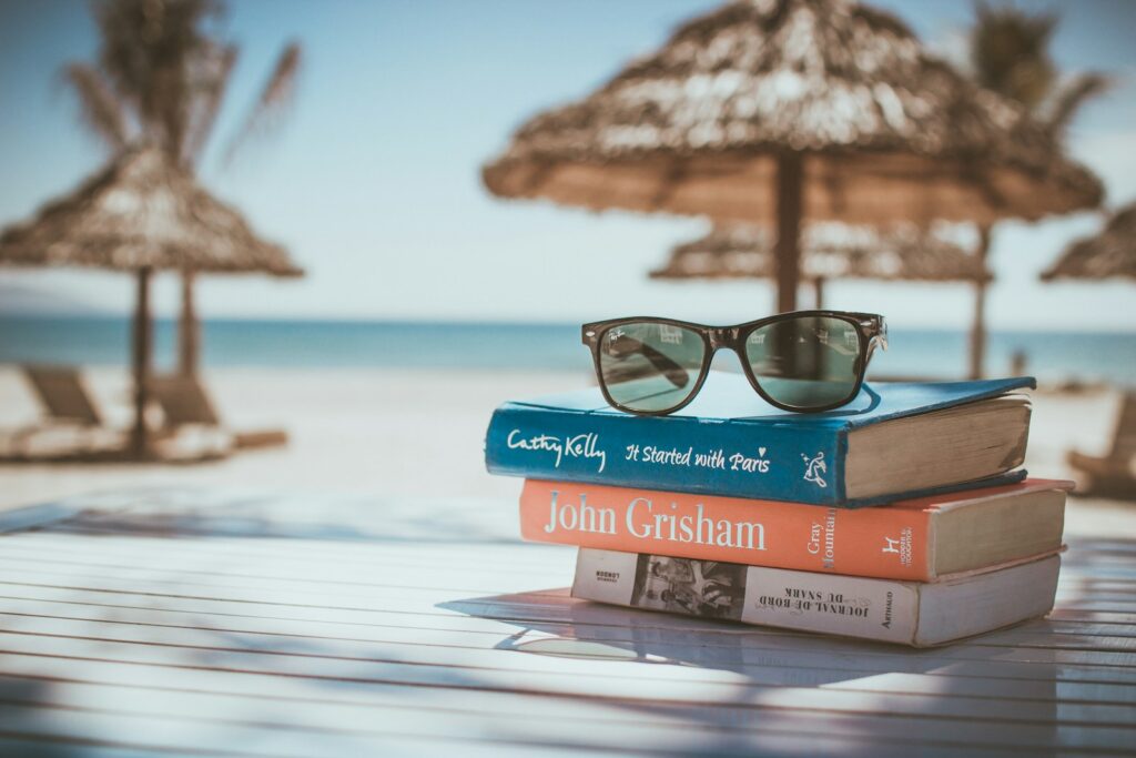 A pile of books with sunglasses on top on a table by a beach.