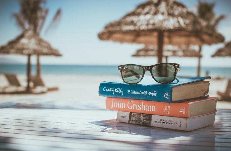 A pile of books with sunglasses on top on a table by a beach.