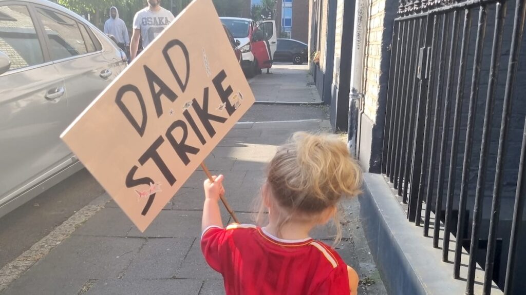 A child holds a Dad Strike placard while walking down a street.