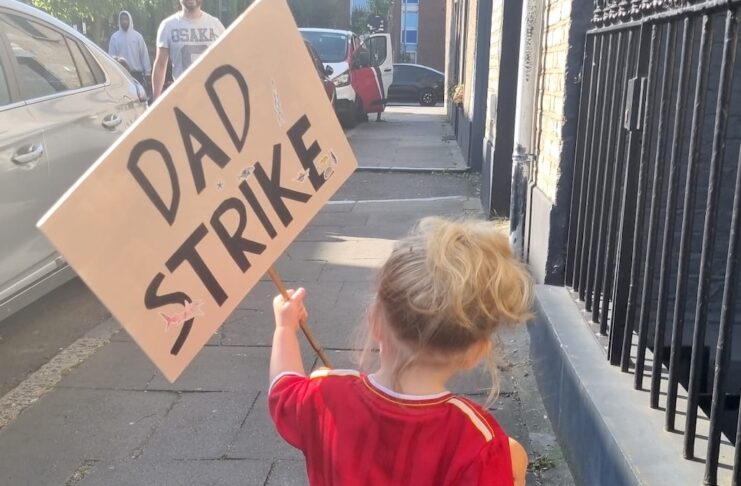 A child holds a Dad Strike placard while walking down a street.