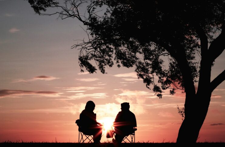 Two people sitting on chairs and near a tree during sunset.