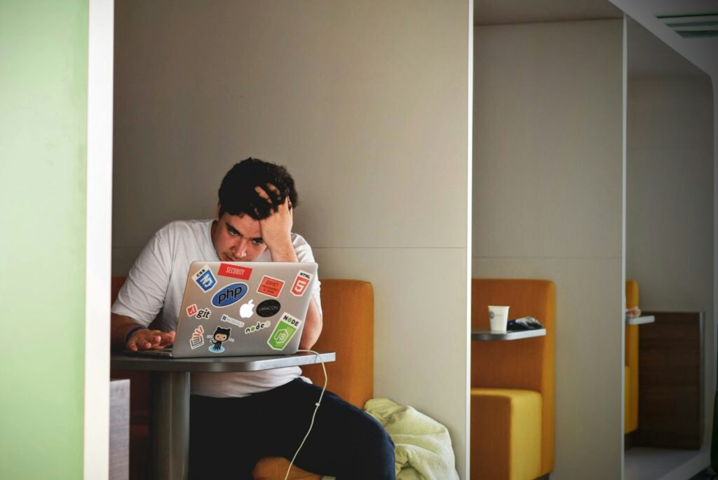 A stressed looking young man at a table with a laptop.