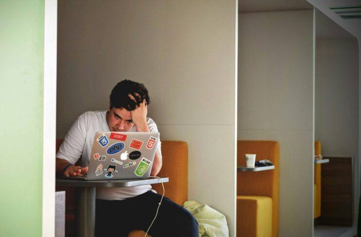 A stressed looking young man at a table with a laptop.