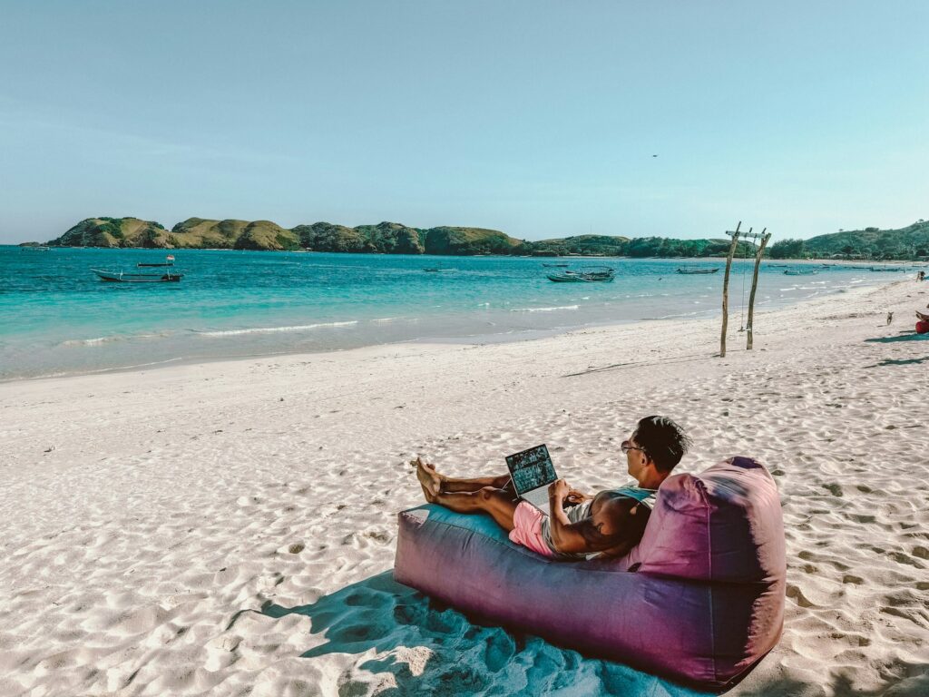 A man sitting on a bean bag on the beach while using a laptop.