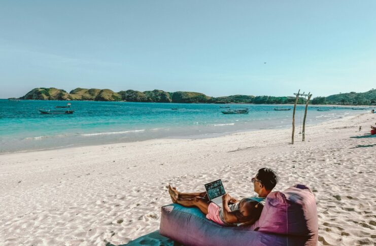 A man sitting on a bean bag on the beach while using a laptop.