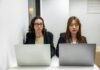 Accent Bias Still Shapes Workplace Confidence and Careers Two female staff sitting at laptops and looking at each other as if there's a conflict between them.