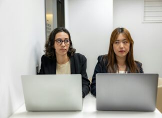 Accent Bias Still Shapes Workplace Confidence and Careers Two female staff sitting at laptops and looking at each other as if there's a conflict between them.