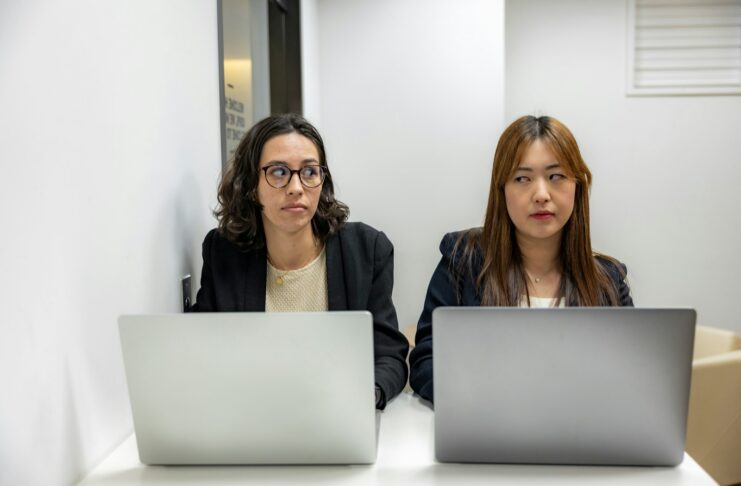 Two female staff sitting at laptops and looking at each other as if there's a conflict between them.