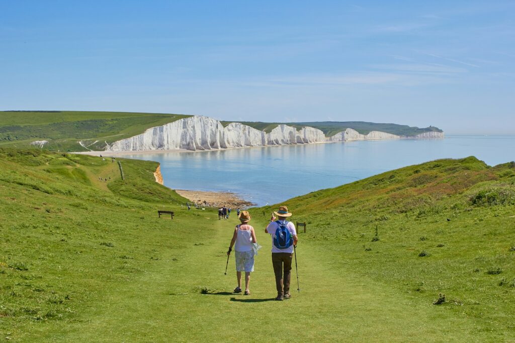 An elderly woman and man walk along a path down to the sea and cliffs on a sunny day.