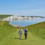 Rising Retirement Age: Keeping Active and Healthy to Support a Longer Career An elderly woman and man walk along a path down to the sea and cliffs on a sunny day.