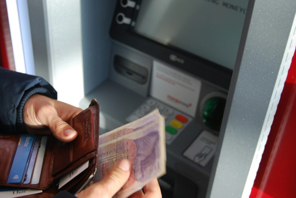 A person holding brown leather wallet and banknotes at an ATM.
