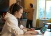 Younger Workers Expect Higher Retirement Income as Anxiety Over Future Finances Grows Young woman with dreadlocks works on a laptop in a casual kitchen environment.