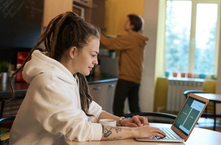 Young woman with dreadlocks works on a laptop in a casual kitchen environment.