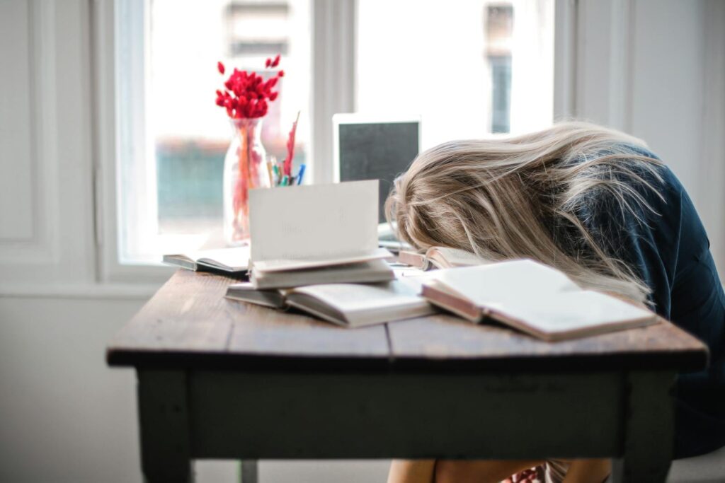 Blonde woman slumped over desk with books, showing burnout.
