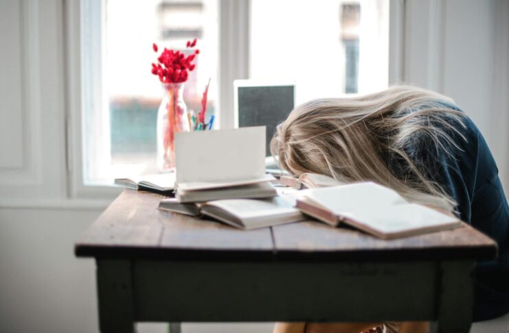 Blonde woman slumped over desk with books, showing burnout.