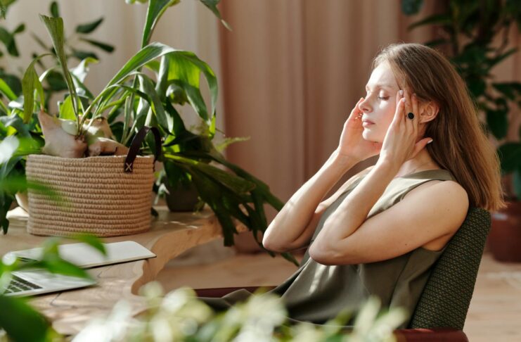 A woman relaxes, massaging her temples among indoor plants in a bright, sunlit room.
