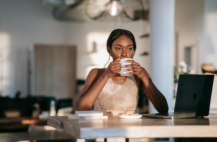A stylish woman drinking coffee and working on a laptop indoors, creating a relaxed atmosphere.