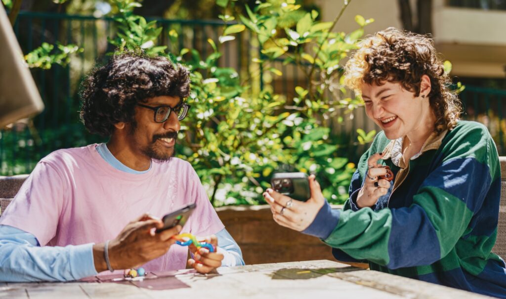 Two autistic friends sitting outside using stim toys and laughing while using phones.