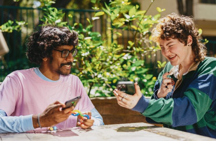Two autistic friends sitting outside using stim toys and laughing while using phones.