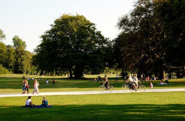 People walking and cycling in a park during a sunny summer's day while others sit on the grass.