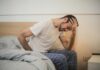 UK Workforce at ‘Breaking Point’ as Over Half Report Jobs are Harming Long Term Health A man in pajamas holding his head and sitting on a bed.