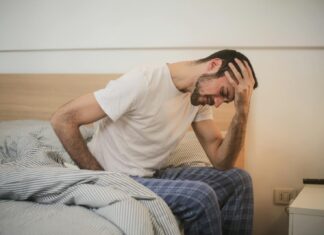 UK Workforce at ‘Breaking Point’ as Over Half Report Jobs are Harming Long Term Health A man in pajamas holding his head and sitting on a bed.
