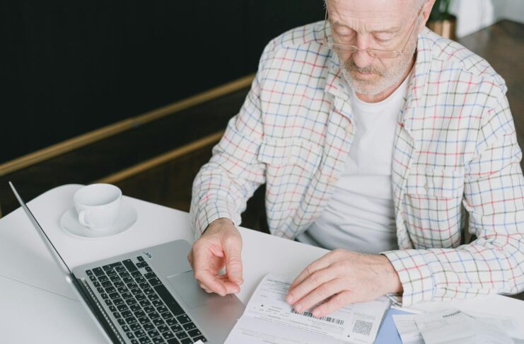 Elderly man with eyeglasses reviewing documents at a laptop. Indoor setting with natural light.