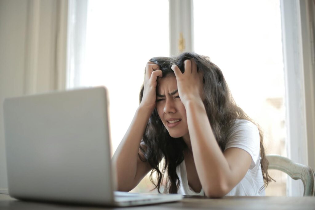 A woman sits in front of a laptop with her hands on her head, looking stressed.