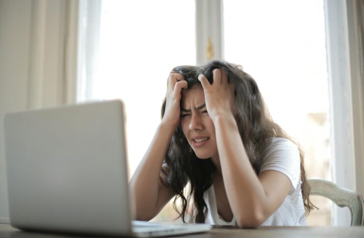 Photo by Andrea Piacquadio A woman sits in front of a laptop with her hands on her head, looking stressed.