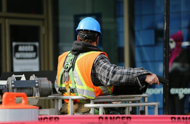 A construction worker wearing a blue helmet and safety vest operates equipment at an urban construction site.