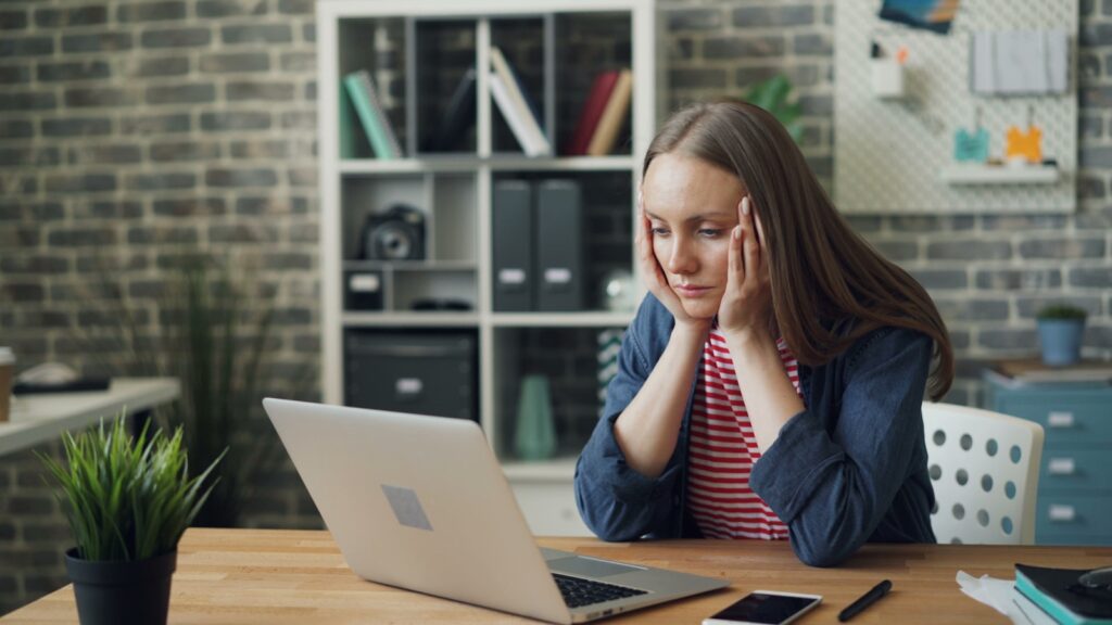 A woman sitting with a laptop in front of her, with her head in her hands, looking overworked.